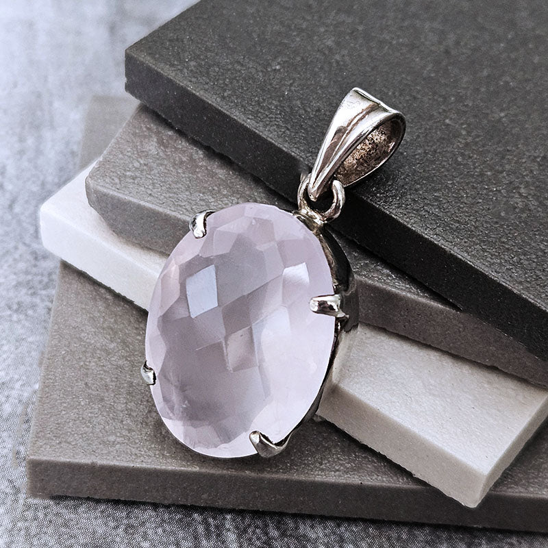 An oval-cut pale pink gemstone pendant in a polished silver-toned bezel setting, displayed on stacked gray and white stone slabs.
