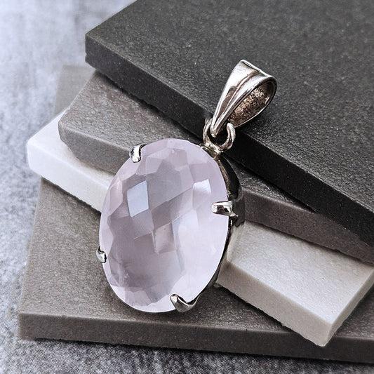 An oval-cut pale pink gemstone pendant in a polished silver-toned bezel setting, displayed on stacked gray and white stone slabs.