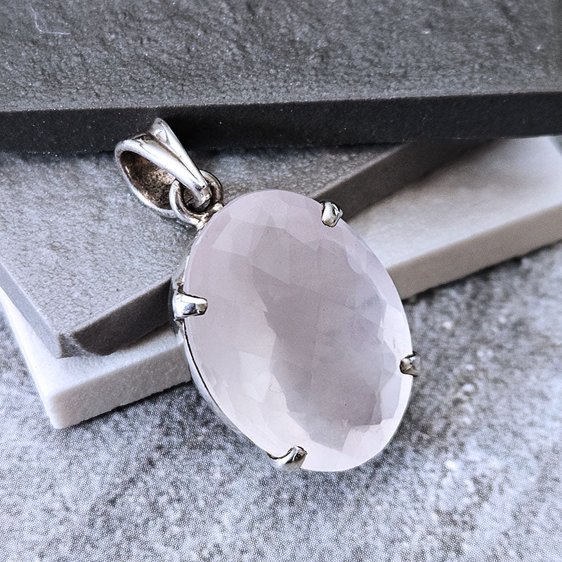 An oval-cut pale pink faceted gemstone pendant in a silver-toned prong setting, displayed on a textured gray surface with dark gray stone blocks.