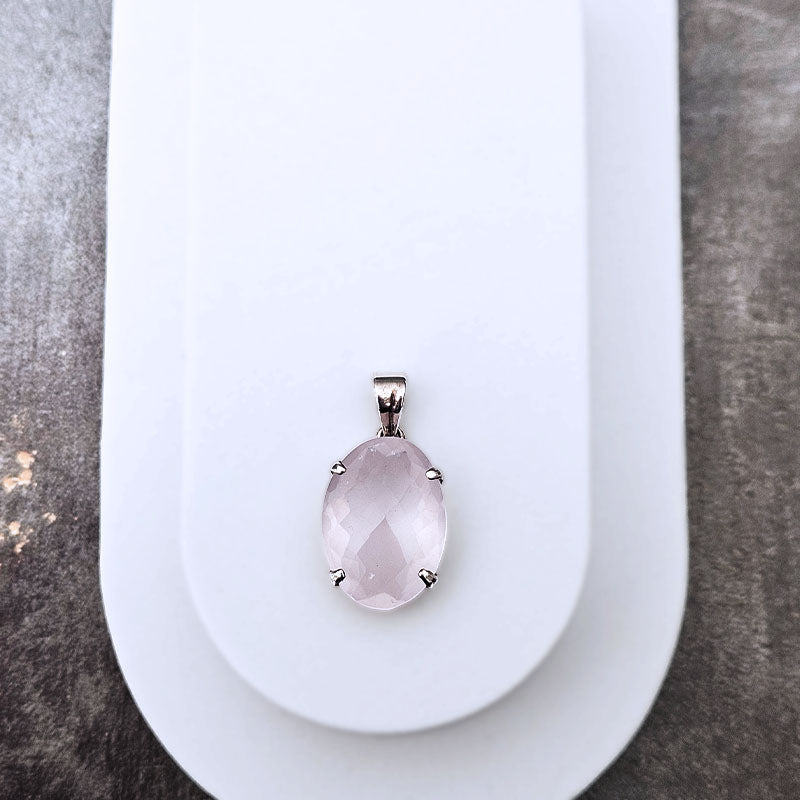 An oval rose quartz pendant in a silver four-prong setting, displayed on a white plastic jewelry stand against a textured gray background.