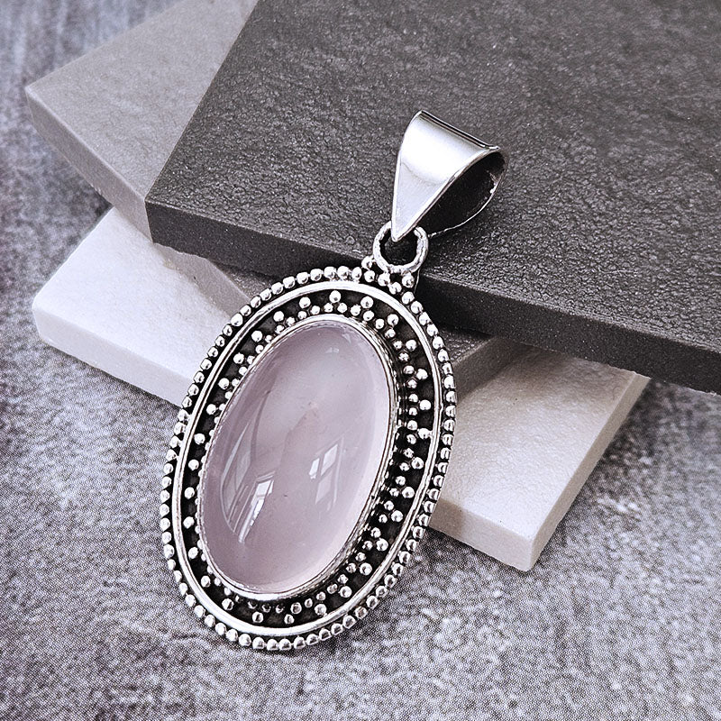 An oval pendant featuring a pale pink cabochon gemstone surrounded by a silver-tone beaded border, displayed on stacked gray and white stone blocks.