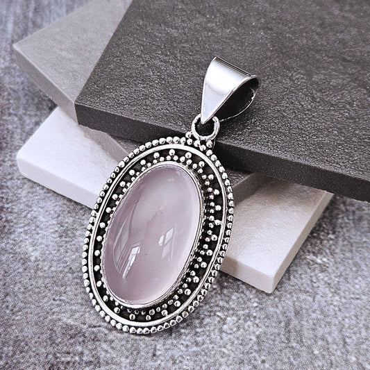An oval pendant featuring a pale pink cabochon gemstone surrounded by a silver-tone beaded border, displayed on stacked gray and white stone blocks.