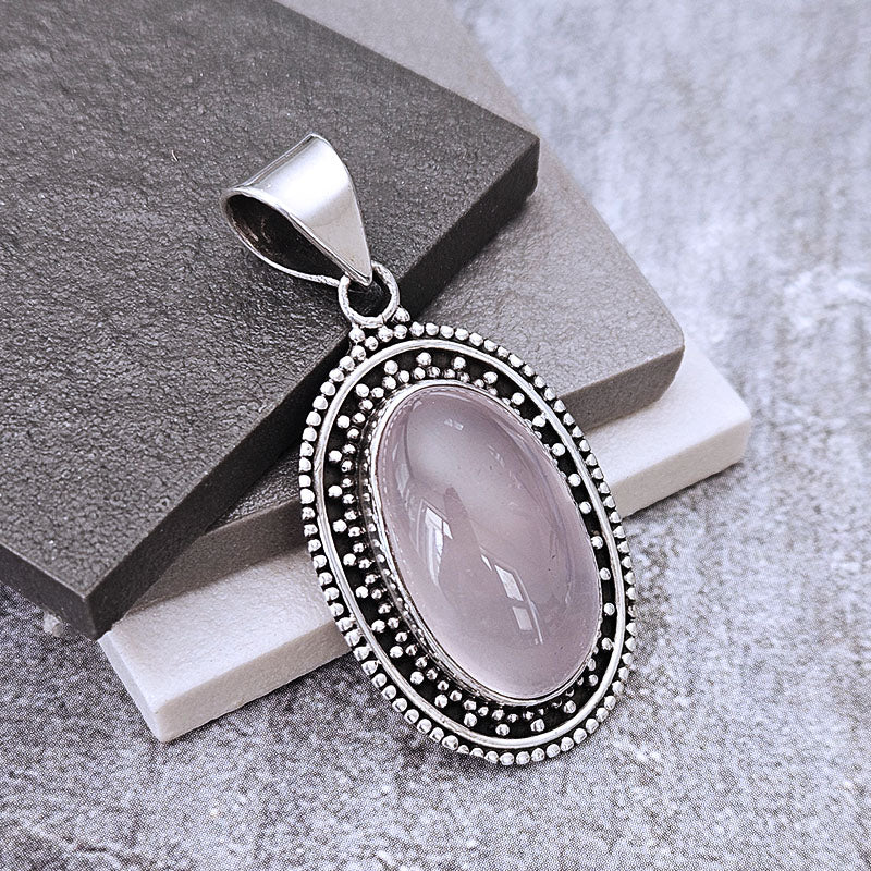 An oval pendant featuring a smooth, pale pinkish-lavender cabochon stone surrounded by a silver-tone beaded border, displayed on a dark gray textured block over a white block against a gray background.