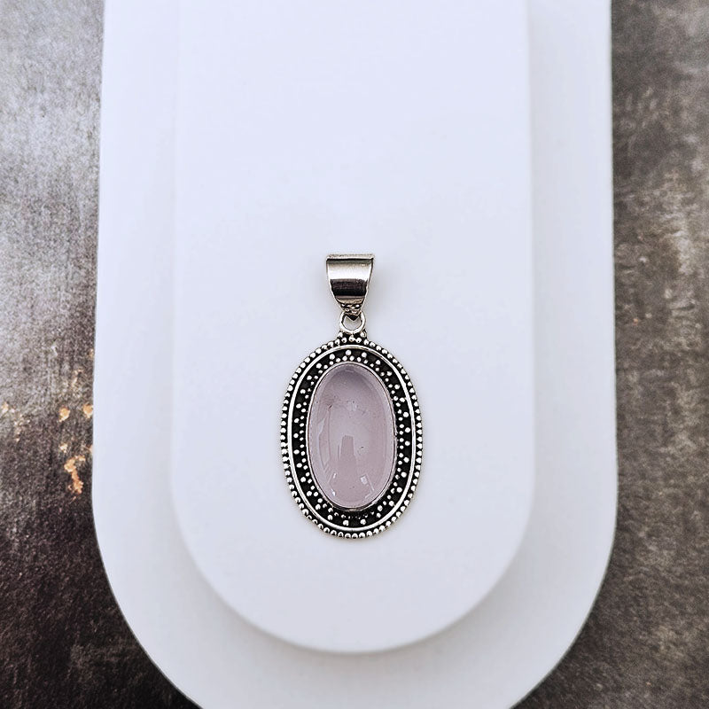 An oval pendant featuring a pale pink cabochon stone surrounded by a silver-tone beaded border, displayed on a white stand against a textured gray background.