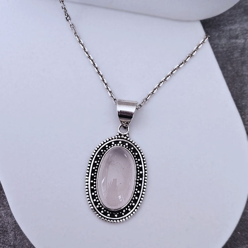 A silver-tone oval pendant necklace featuring a pale pink stone in the center, surrounded by a beaded silver border, displayed on a white jewelry stand.