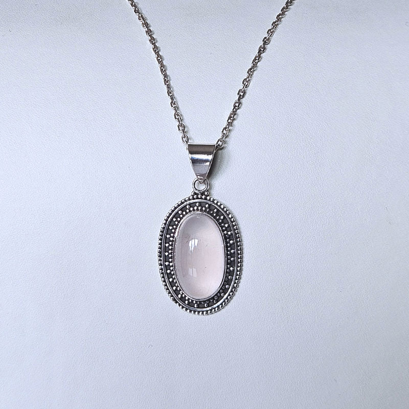 An oval pendant necklace featuring a pale pink cabochon stone surrounded by textured silver borders, suspended from a silver chain against a light gray background.