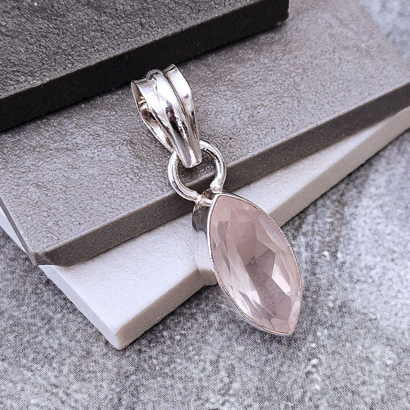 A silver pendant featuring a faceted marquise-cut light pink gemstone, displayed on a textured gray stone surface.