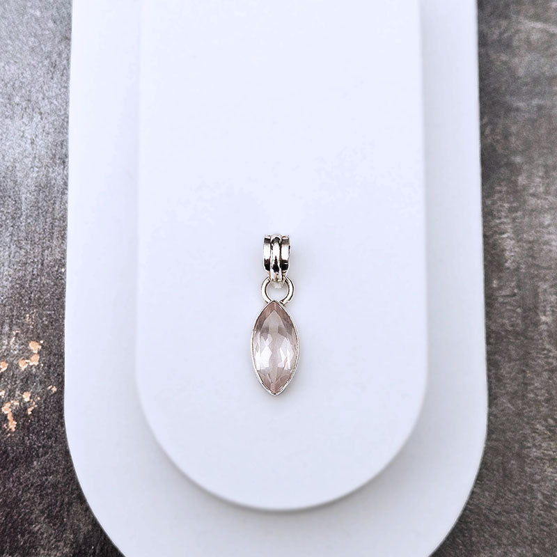 A silver-toned marquise-shaped charm with a faceted pale pink gemstone, displayed on a white card against a textured gray background.