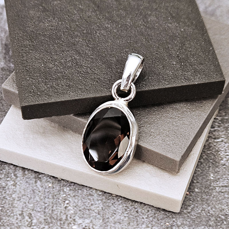 A silver pendant with an oval dark smoky quartz stone, displayed on stacked dark gray and white textured blocks.
