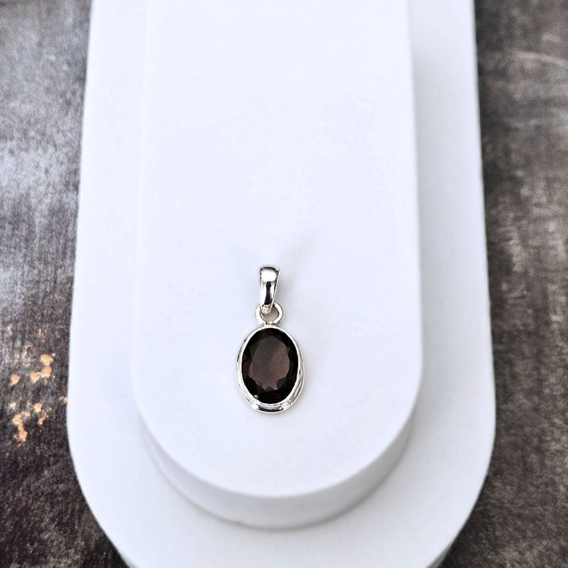 A silver oval pendant with a dark brown smoky gemstone, displayed on a white jewelry stand against a dark textured background.