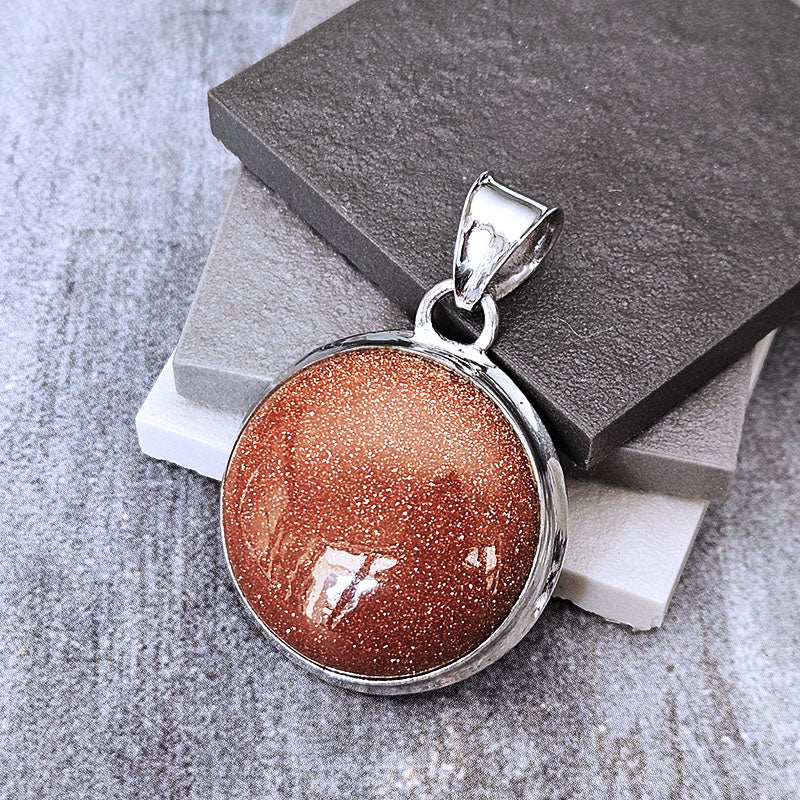 A round pendant with a shimmering coppery-brown glitter stone in a silver bezel, displayed on stacked gray and white stones.