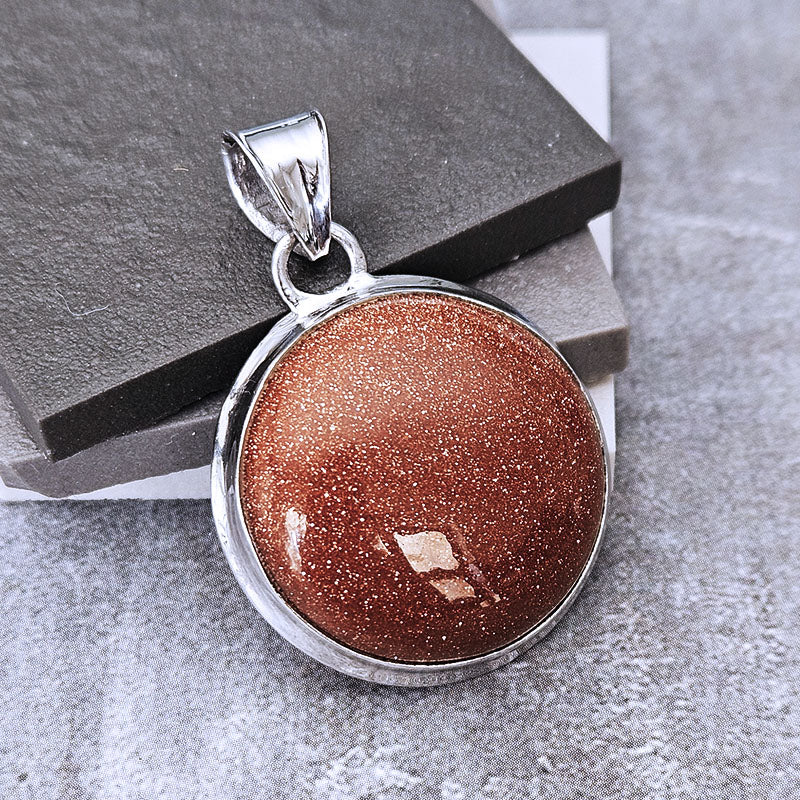 A round silver pendant featuring a shimmering coppery-brown glitter stone, displayed on a dark gray stone block over a textured gray surface.