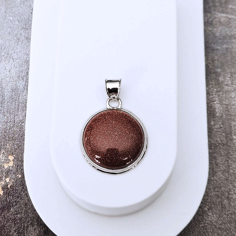 A round silver pendant with a sparkling bronze-brown glitter center, displayed on a white stand against a textured gray background.