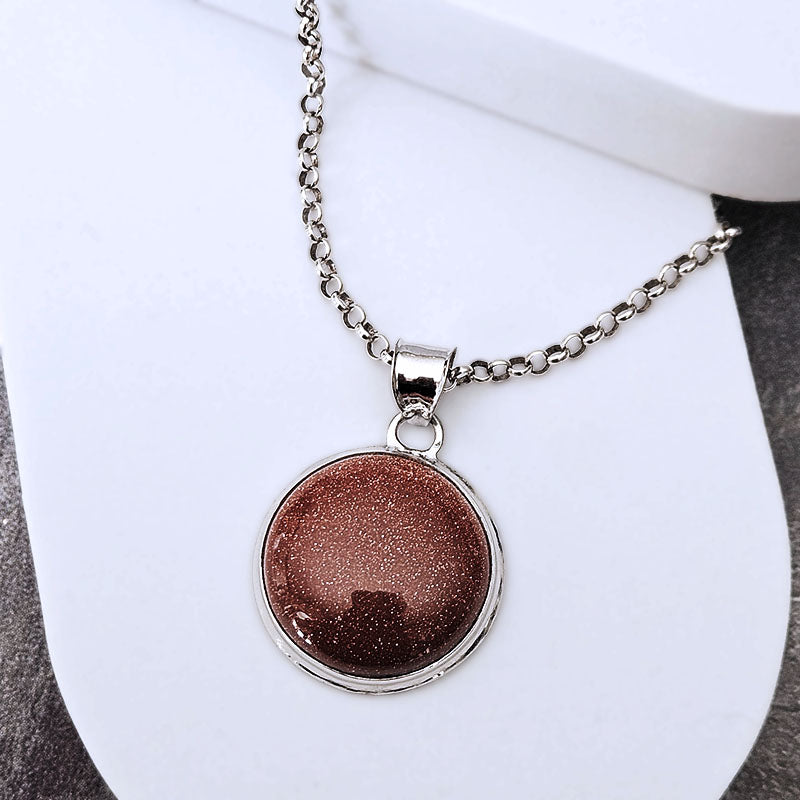 A silver chain necklace with a round pendant featuring a shimmering bronze-brown glitter center, displayed on a white jewelry stand.
