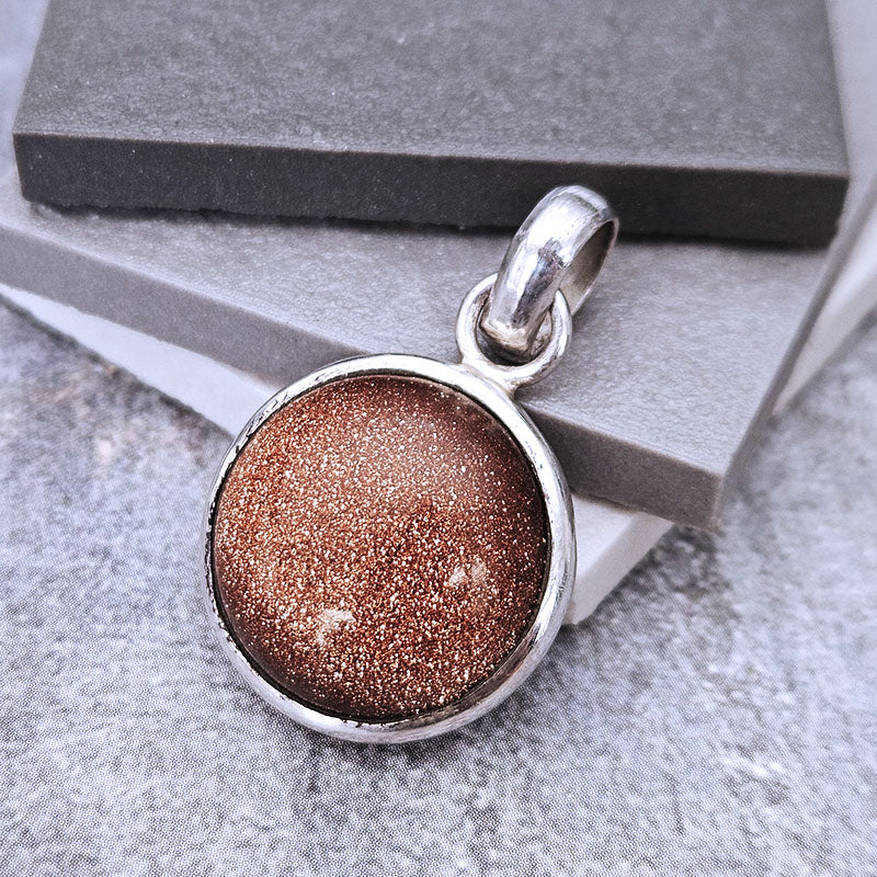 A round silver pendant with a sparkling bronze-brown glitter center, resting on stacked gray display blocks.