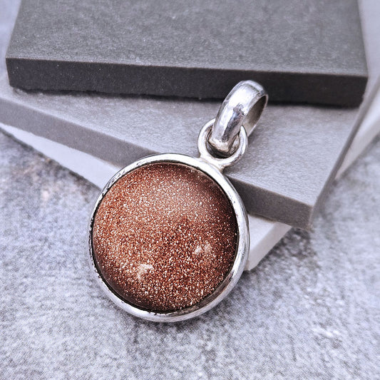 A round silver pendant with a sparkling bronze-brown glitter center, resting on stacked gray display blocks.