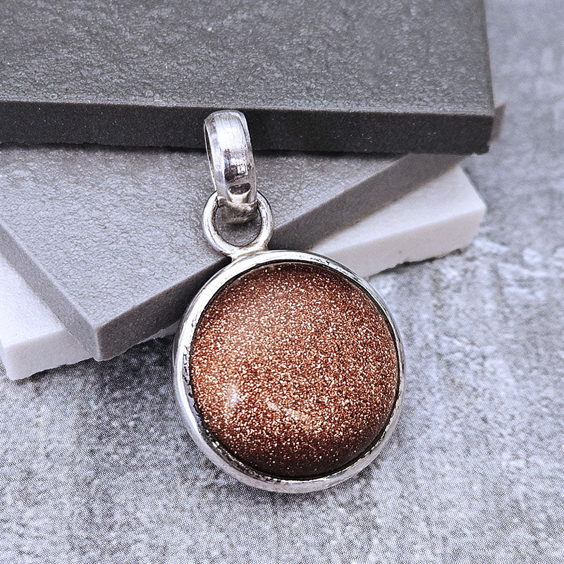 A round silver pendant featuring a sparkling coppery-brown glitter stone, displayed on stacked gray and white stone blocks.