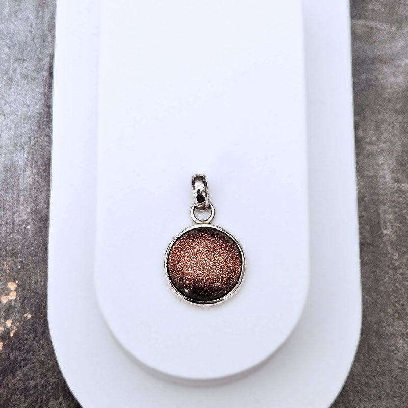 A round metallic pendant with a glittery rose gold finish, set in a silver-toned bezel with a small loop at the top, displayed on a white card over a gray textured surface.