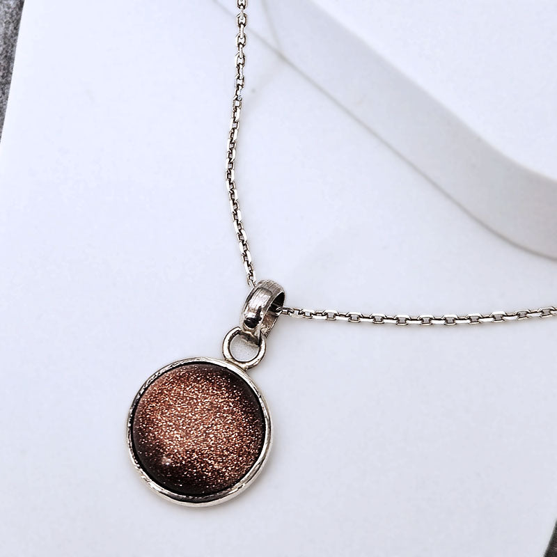 A silver chain necklace with a round pendant featuring a metallic bronze glitter finish, displayed on a clean white surface.