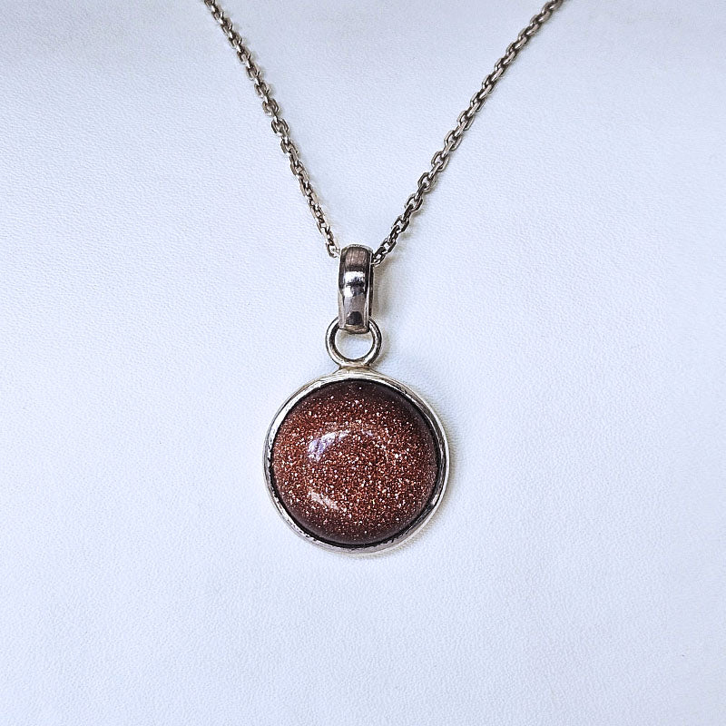 A silver chain necklace with a round bezel pendant containing a sparkling coppery-brown glittery stone, displayed on a light off-white background.