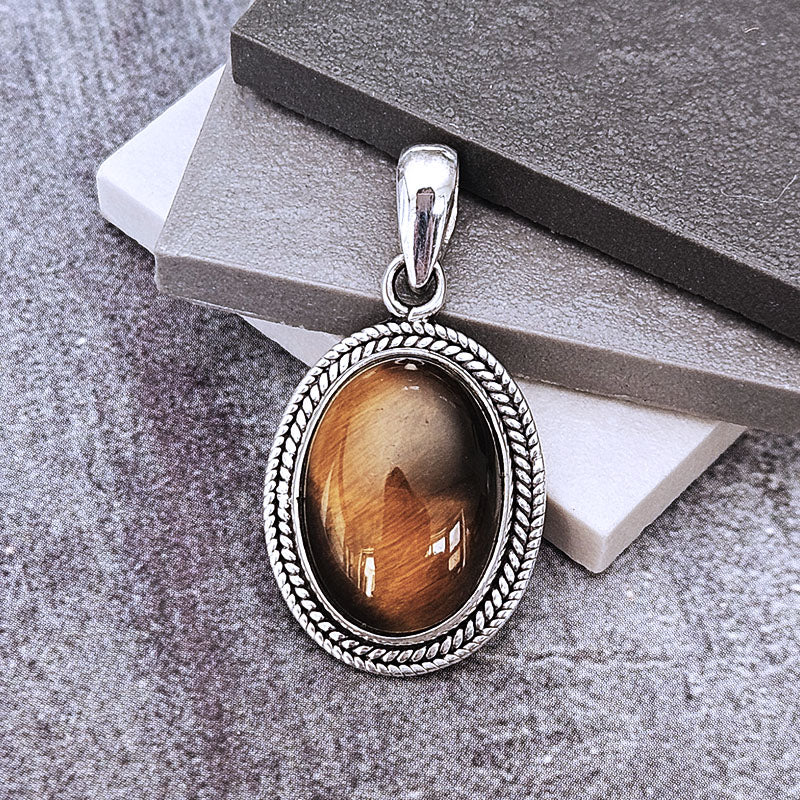 An oval pendant featuring a brown tiger's eye stone in a silver-toned rope-textured bezel, displayed on stacked gray and white blocks.