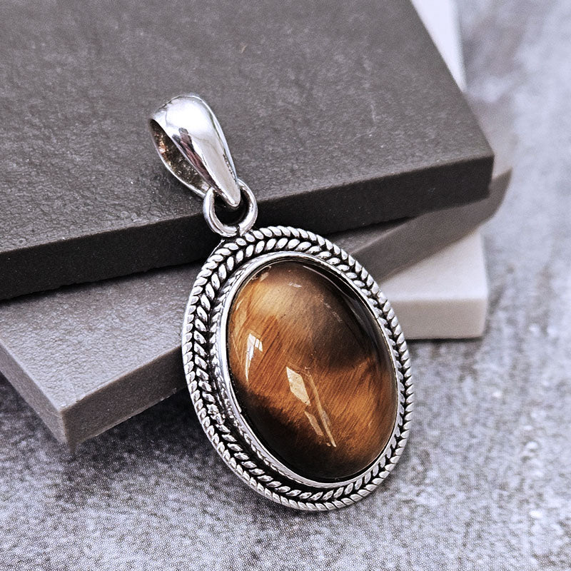A silver-toned oval pendant with a textured rope border and a brown tiger's eye stone, displayed on stacked dark gray and white blocks.