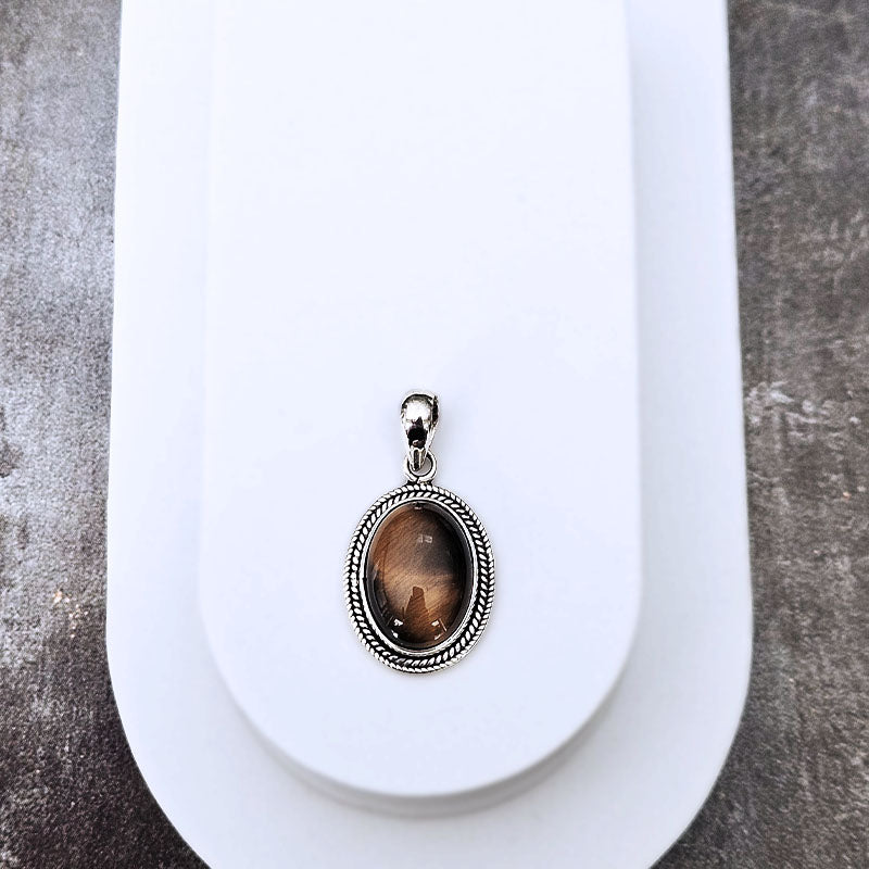 An oval pendant with a silver-toned rope-textured border and a brown cat-eye stone, displayed on a white jewelry stand against a gray textured background.