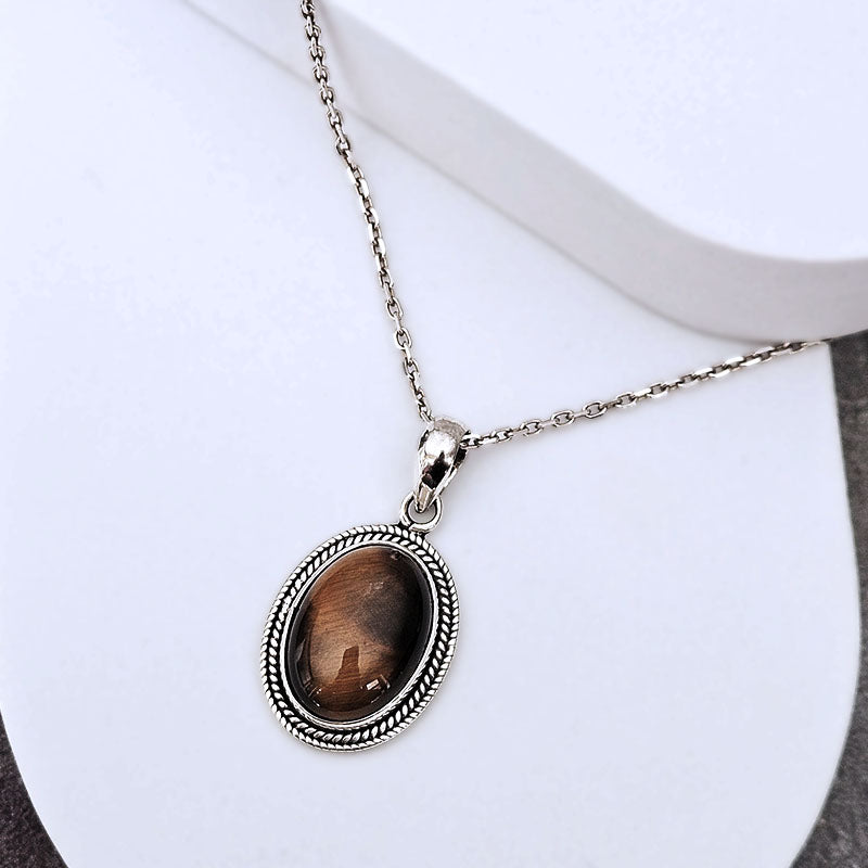 An oval pendant necklace with a brown tiger's eye stone set in a silver-toned rope border, displayed on a white jewelry stand.