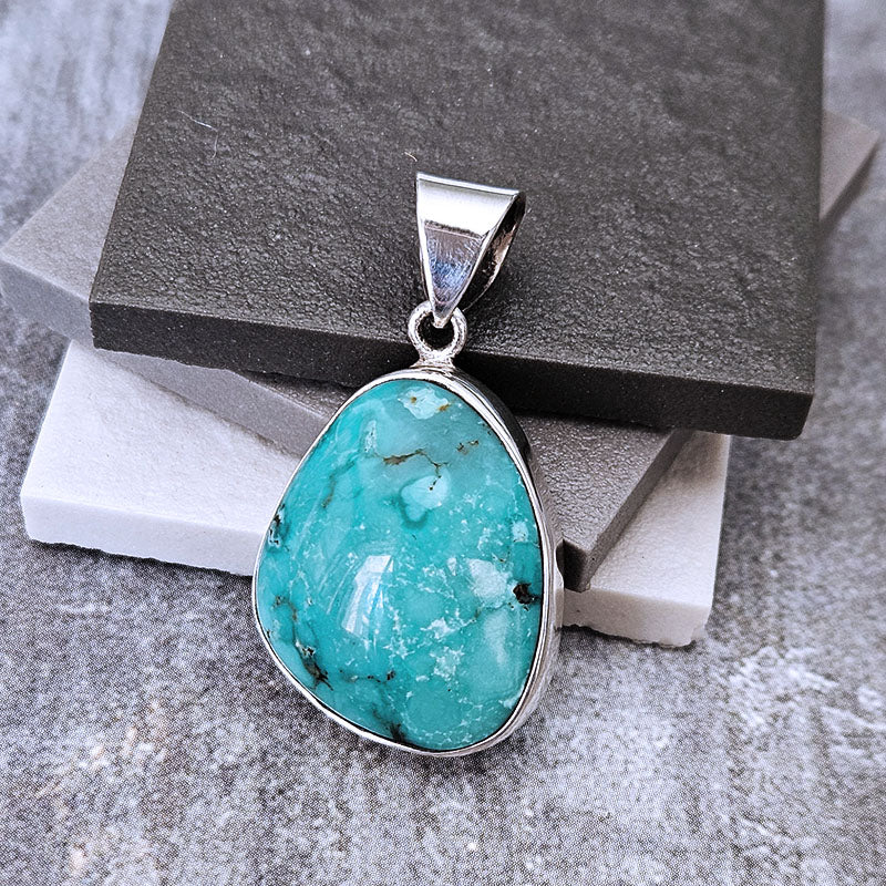 A turquoise-colored stone pendant with a silver bezel setting and bail, displayed on stacked dark and light gray stone blocks over a textured gray surface.