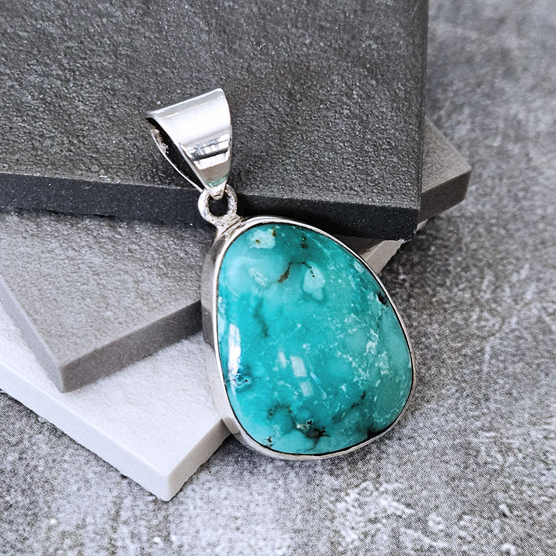 A silver pendant featuring a large, teardrop-shaped turquoise stone with natural dark veining, displayed on stacked gray and white stone blocks.