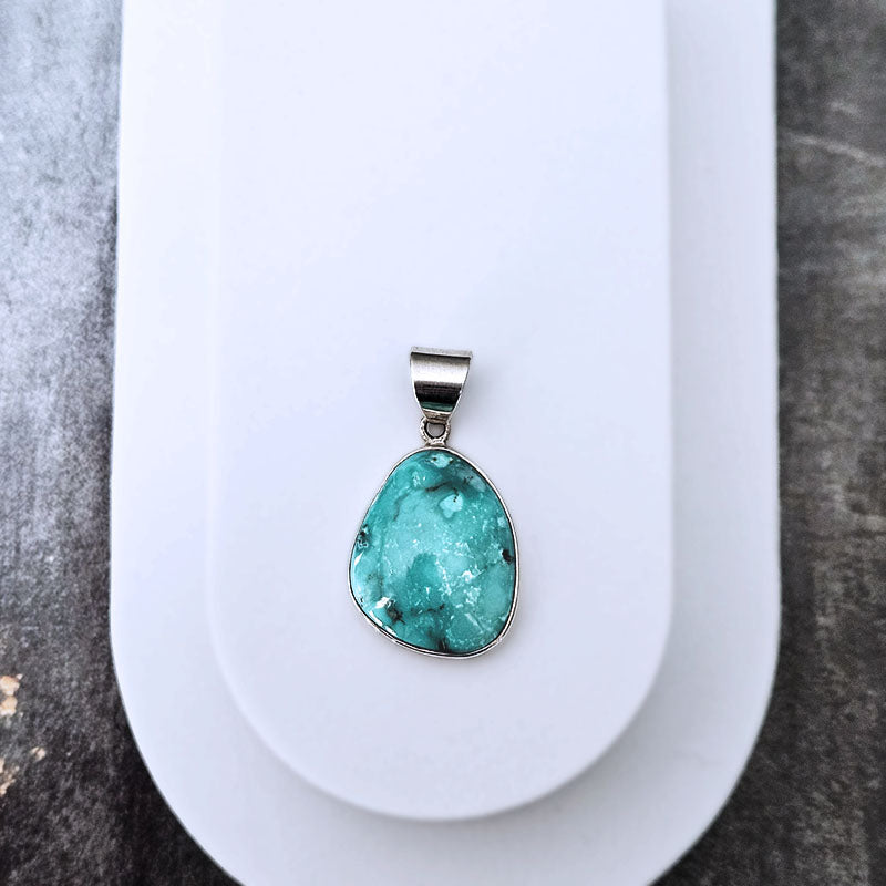 A teardrop-shaped turquoise pendant with a silver bezel setting, displayed on a white card over a dark textured surface.