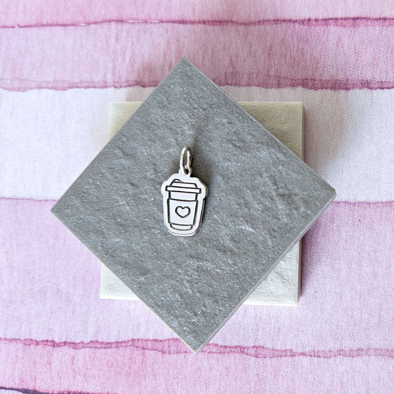 Silver charm of a coffee cup with a lid on a gray card against a pink and white striped background