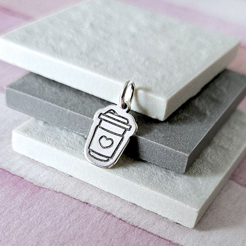 Silver pendant shaped like a coffee cup with a heart on a stone surface