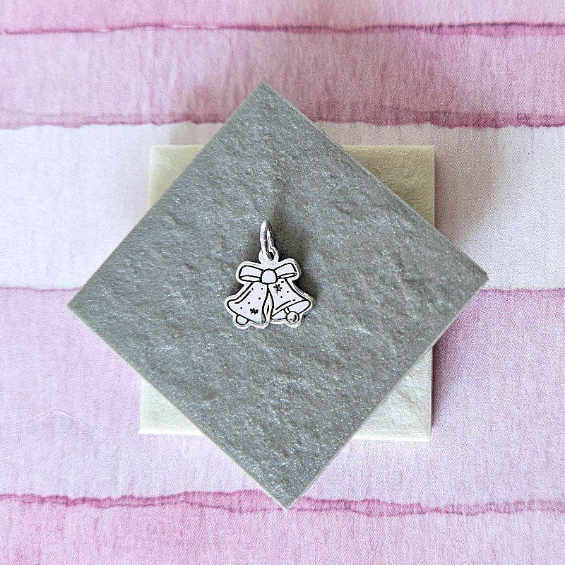 Silver bell-shaped charm with a bow on a gray card against a pink fabric background