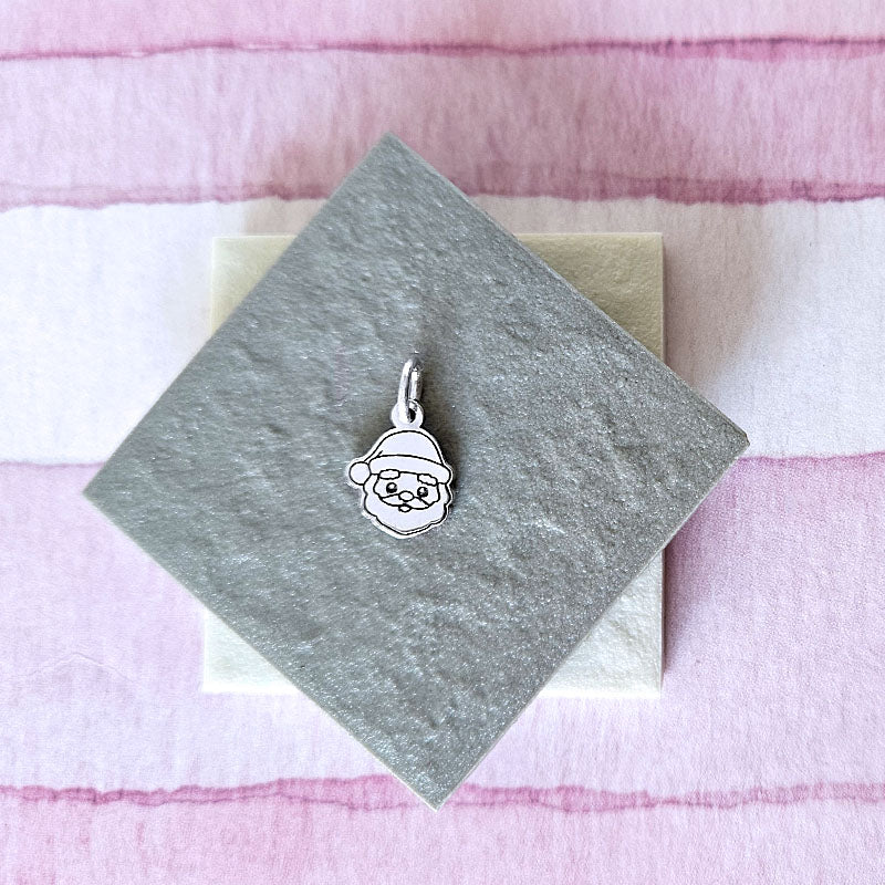 Silver pendant of a face with a Santa hat on a gray card with a pink and white striped background