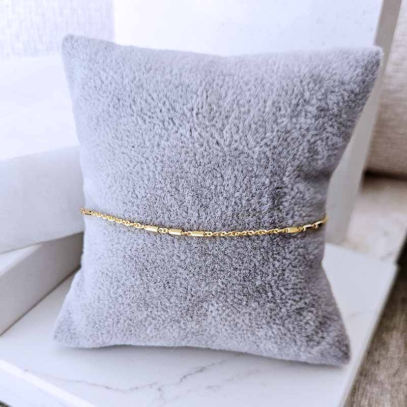 A delicate gold chain bracelet displayed on a soft, textured gray pillow, set against a clean white marble surface.