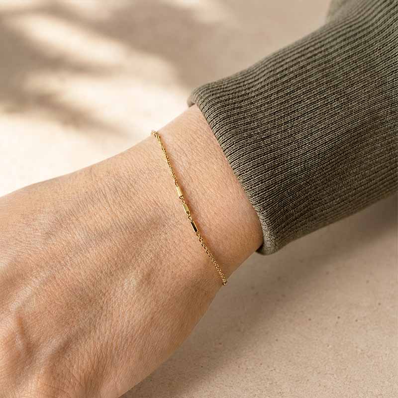 A delicate gold chain bracelet worn on a person's wrist, shown against a neutral beige background with soft shadows and a ribbed olive-gray sleeve visible.