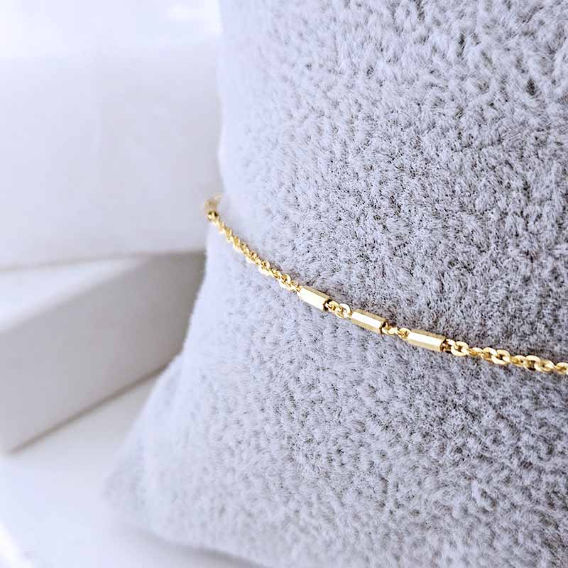 A delicate gold chain bracelet with polished cylindrical links, draped over a textured light gray fabric display pillow.