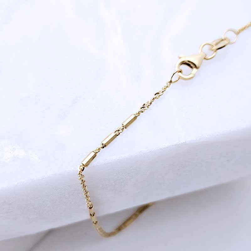 A close-up of a gold chain bracelet with a lobster clasp, resting on a white textured surface.
