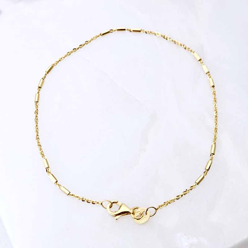 A delicate gold chain bracelet with cylindrical links and a lobster clasp, displayed on a white marble surface.