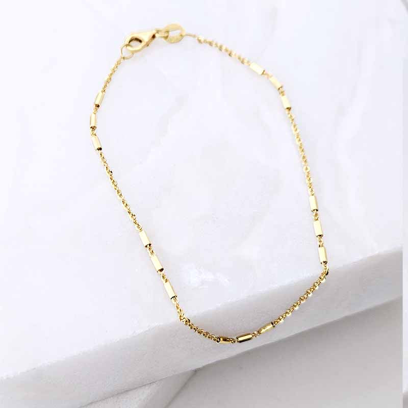 A delicate gold chain bracelet with alternating small round beads and polished rectangular links, displayed on a white marble surface.