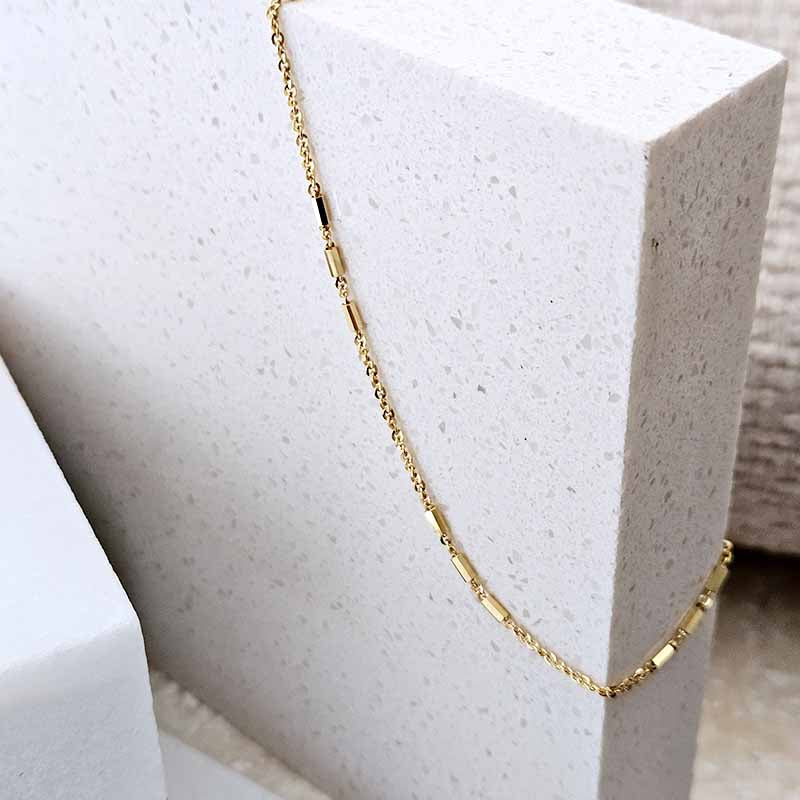 A delicate gold chain necklace with polished rectangular links, draped over a textured white stone display block.