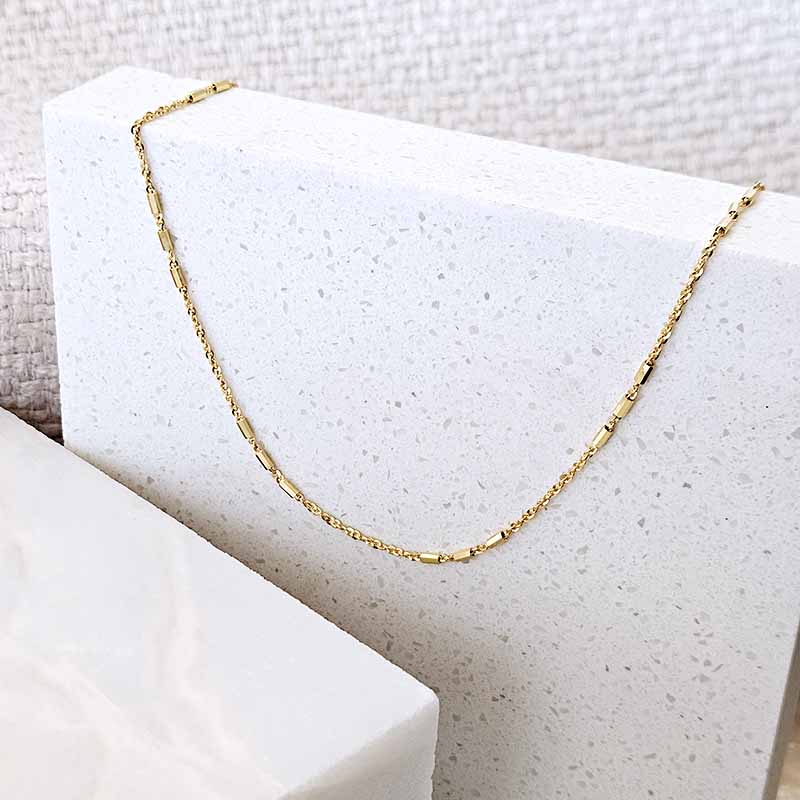 A delicate gold chain necklace draped over a white speckled display block on a neutral textured background.