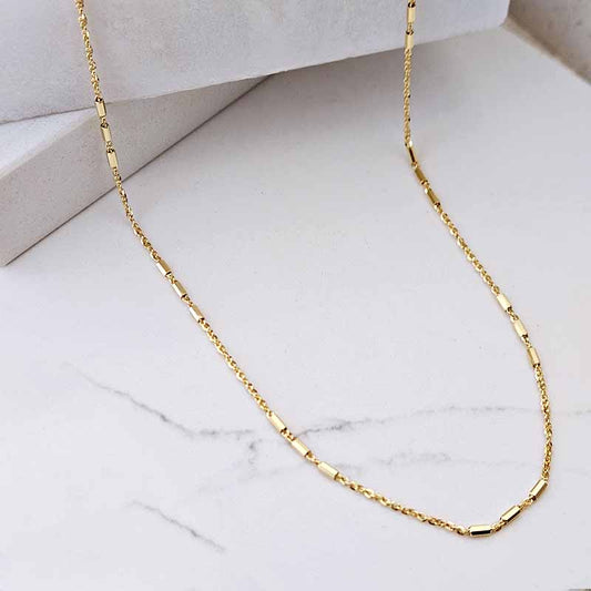 A gold chain necklace with a modern segmented link design, displayed on a white marble surface with a white geometric block in the background.