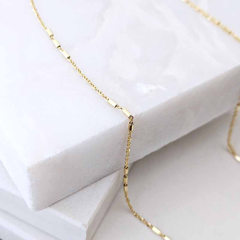 A delicate gold chain necklace with a central barbell motif draped over stacked white marble blocks.