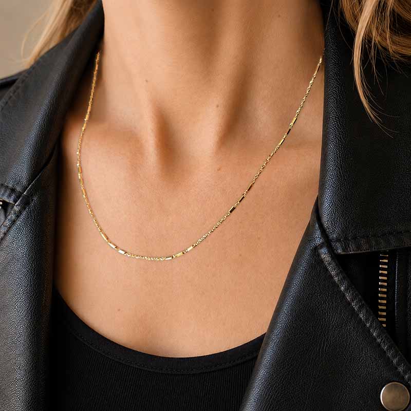 A close-up of a person's neck and chest wearing a delicate gold chain necklace, styled with a black leather jacket and black top.