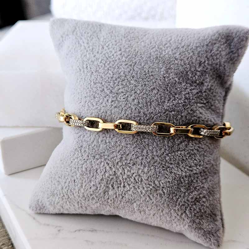 A gold chain bracelet with blank links and clear crystal accents, displayed on a gray plush pillow.