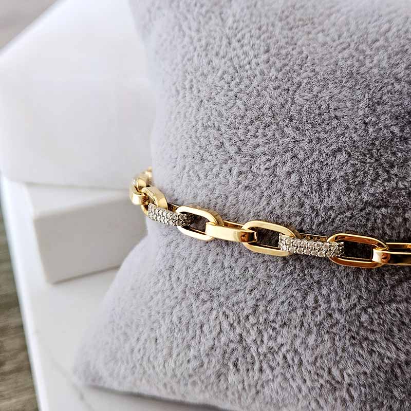 A gold chain bracelet with textured pavé details, displayed on a soft gray textured pillow.