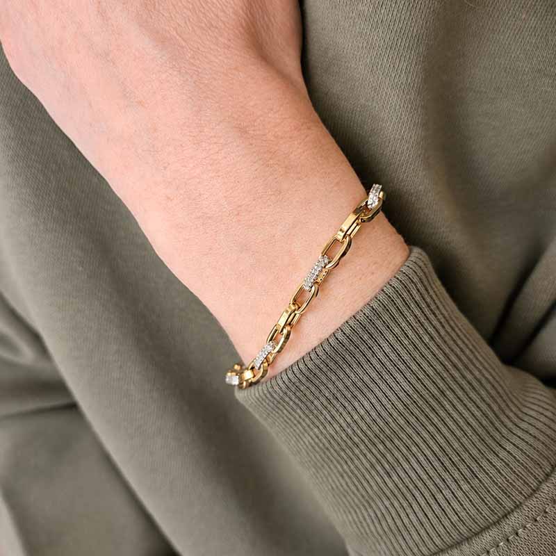 A close-up of a gold chain bracelet with clear gemstone accents worn on a person's wrist, paired with an olive green ribbed knit sweater.