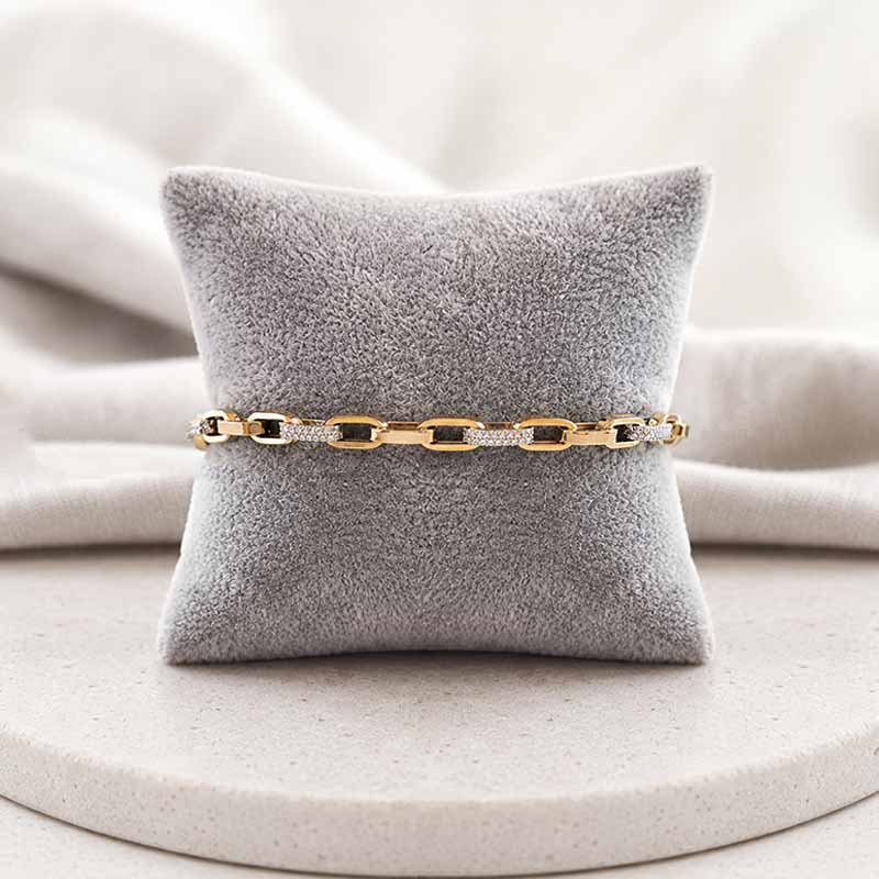 A gold chain bracelet with alternating clear crystal and blank rectangular links, displayed on a gray velvet jewelry pillow over a light beige surface.